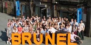 An image of the Brunel graduates standing in front of an orange Brunel sign in front of Bargehouse.
