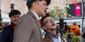 Two men sing into a microphone. Both are wearing brown blazers, and one is wearing a brown flat cap.