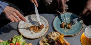 A table is laden with plates of food. Two people's hands cut food on plates