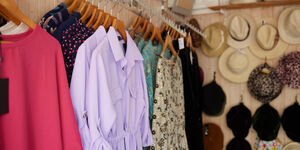 Colourful clothing hang on wooden hangers, behind which is a wall of hats