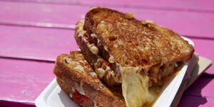 A toasted sandwich filled with melted cheese in a white carton, on a pink wooden bench