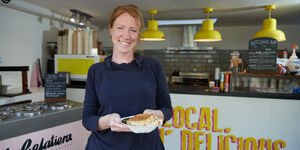 A woman stands in a cafe holding a white carton containing a toasted cheese sandwich