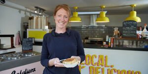 A woman stands in a cafe holding a carton containing a sandwich