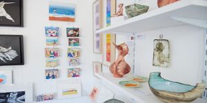 The interior of a shop with white walls hung with paintings and shelves with ceramics and glass items