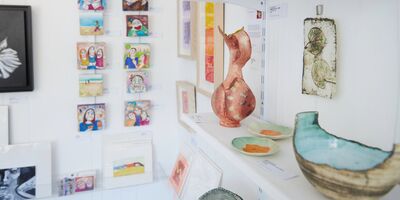 A photo of a white-walled gallery with shelves of ceramics and walls hung with paintings