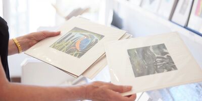 A photograph of a person holding two prints in white card frames. Only the person's hands and arms are visible.