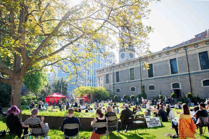 people sitting on the grass and on chairs, watching performers under a red marquee